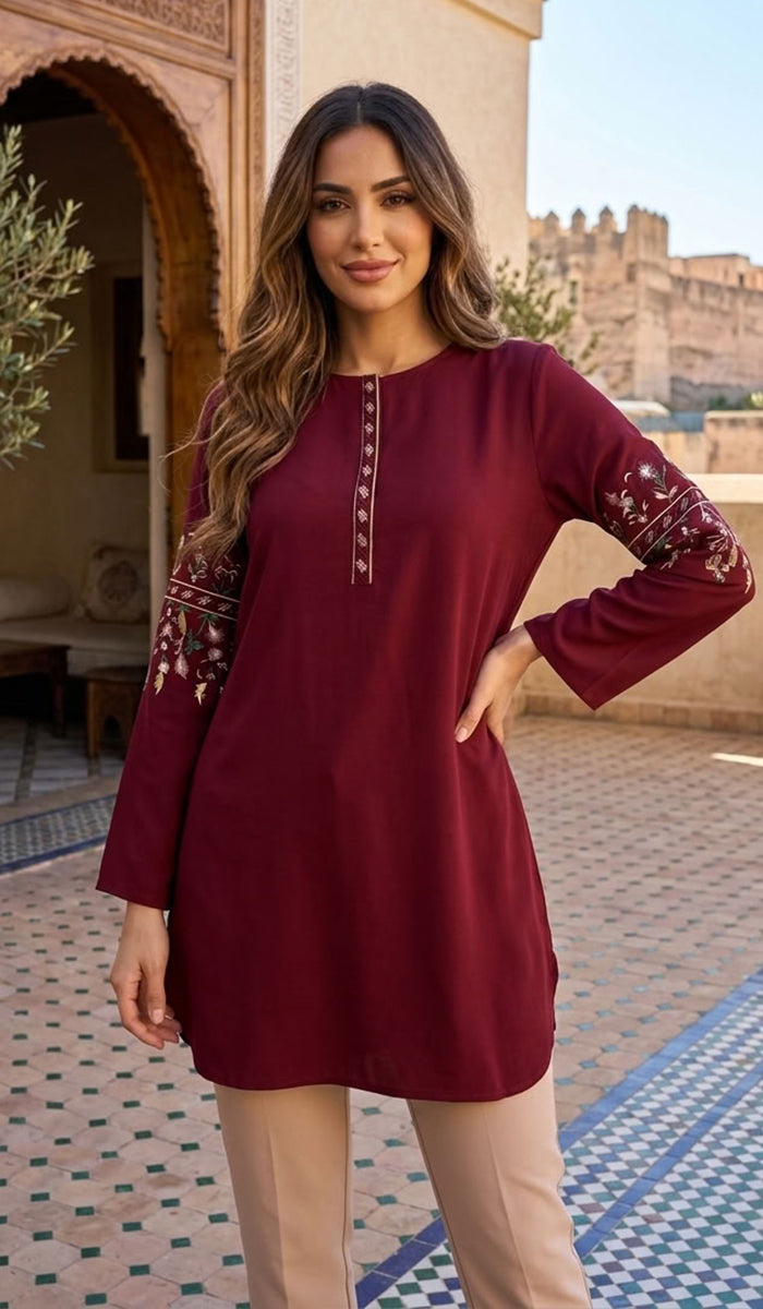 A woman modeling a long-sleeve maroon tunic top with embroidered details on the sleeves and neckline, paired with beige trousers, set against a Moroccan-style courtyard background.