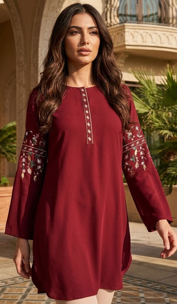 A woman wearing a long-sleeved maroon tunic dress with white and gold embroidery on the sleeves and a gold button placket, standing in front of a warm-toned building with palm trees.