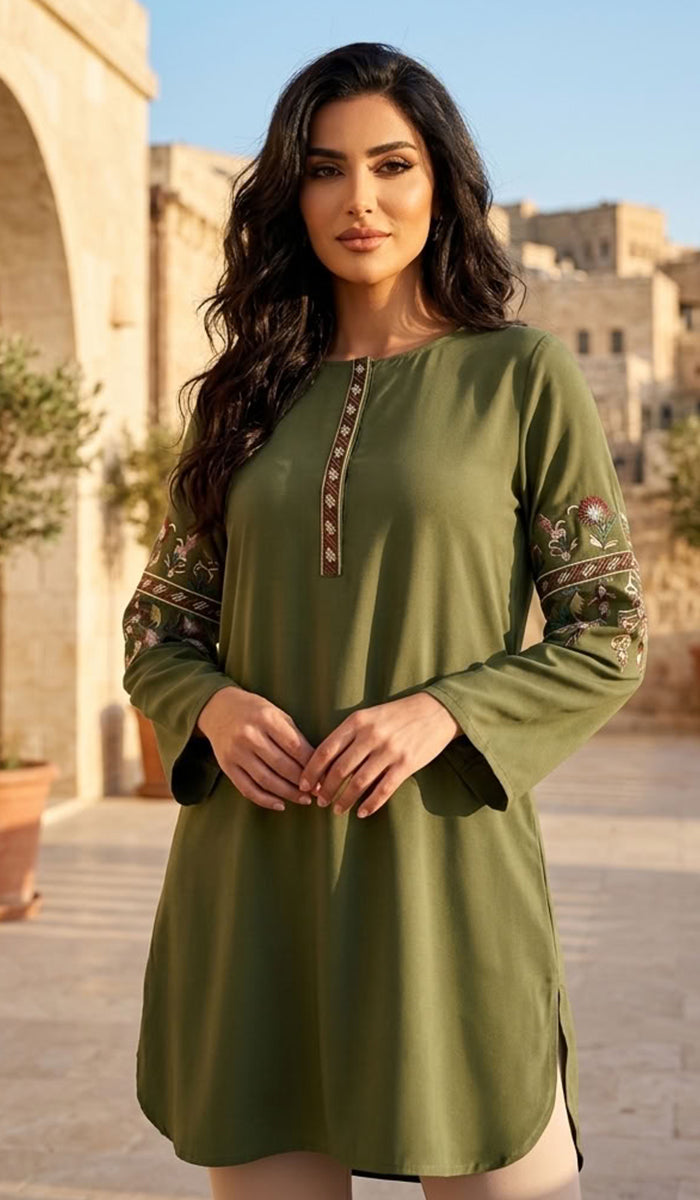 A woman modeling a long-sleeve olive green tunic dress with embroidered details on the sleeves and neckline, set against a warm, sunlit stone courtyard background.