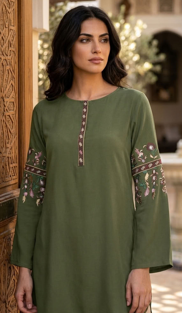 A woman wearing an olive green long-sleeve tunic dress with embroidered details on the neckline and sleeves, standing in front of a carved wooden door.