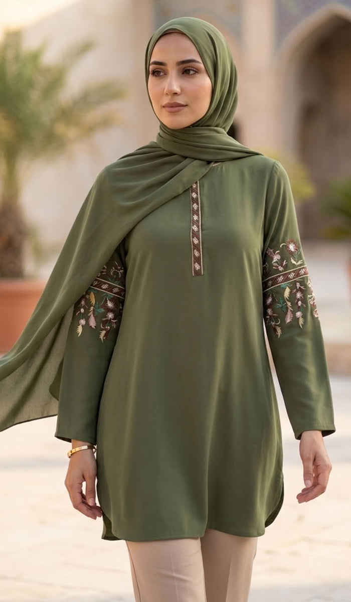 A woman wearing an olive green long-sleeve tunic with floral embroidery on the sleeves and a matching draped hijab, standing in an outdoor architectural setting.