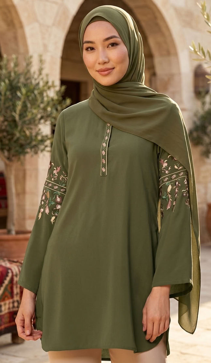 A woman wearing an olive green modest tunic with floral embroidery on the sleeves and a matching hijab, standing in front of a stone archway.