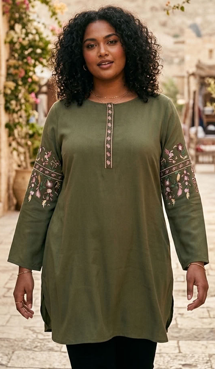 A woman wearing an olive green tunic top with floral embroidery on the sleeves and a decorative neckline, standing in an outdoor street setting.