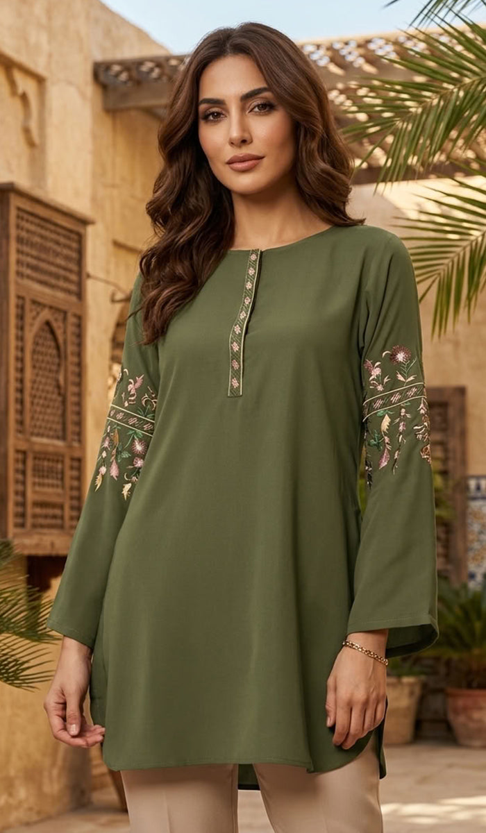 A woman wearing an olive green long-sleeve tunic with floral embroidery on the sleeves and a decorative neckline, standing in front of a warm-toned traditional building with palm leaves.
