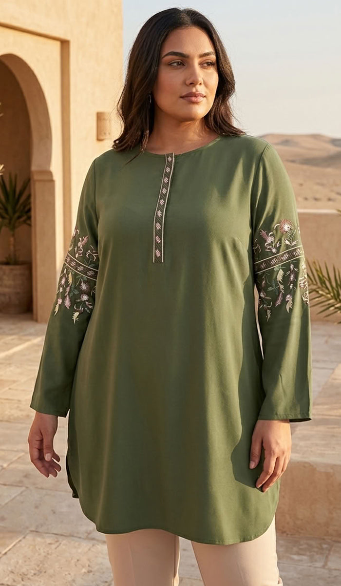 A woman modeling a sage green tunic with floral embroidery on the sleeves and a decorative vertical trim down the center front, set against a warm desert architectural background.