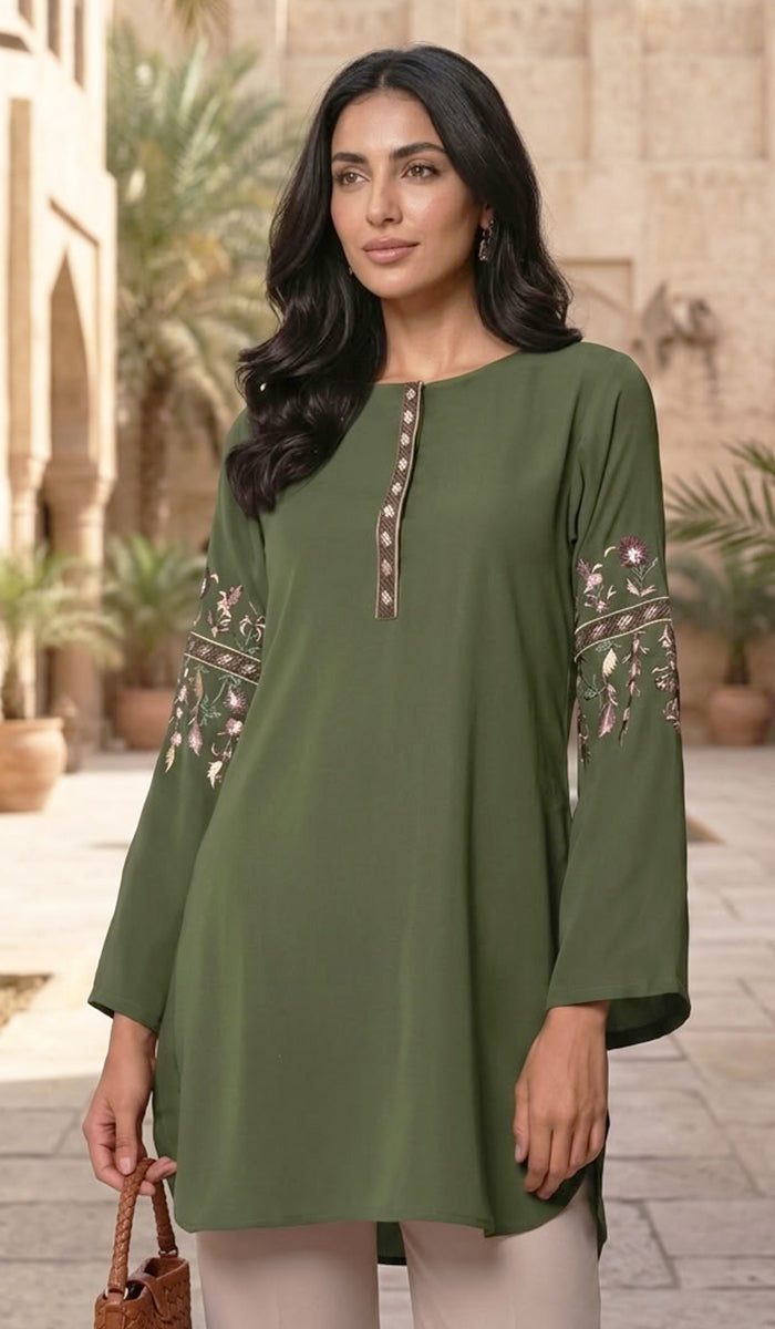 A woman wearing an olive green tunic top with floral embroidery on the sleeves and a decorative button placket, standing in an outdoor courtyard setting.
