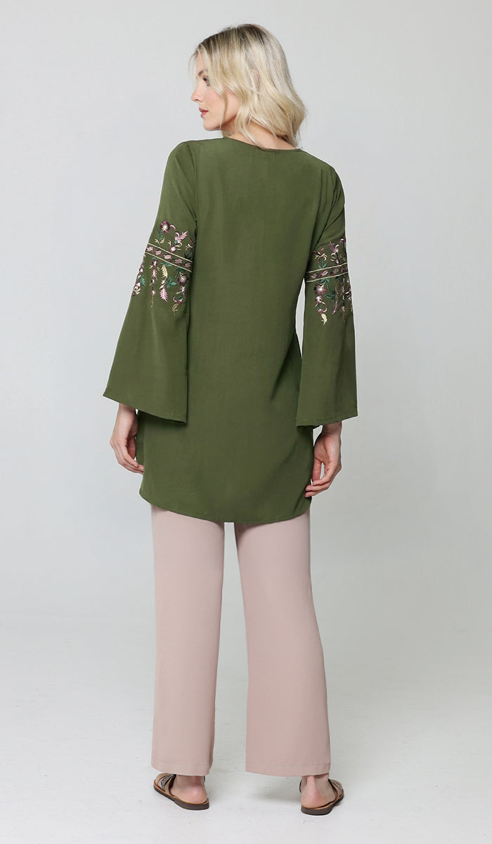 A woman shown from the back wearing an olive green tunic with floral embroidered sleeves and light pink wide-leg pants, standing against a light gray studio background.