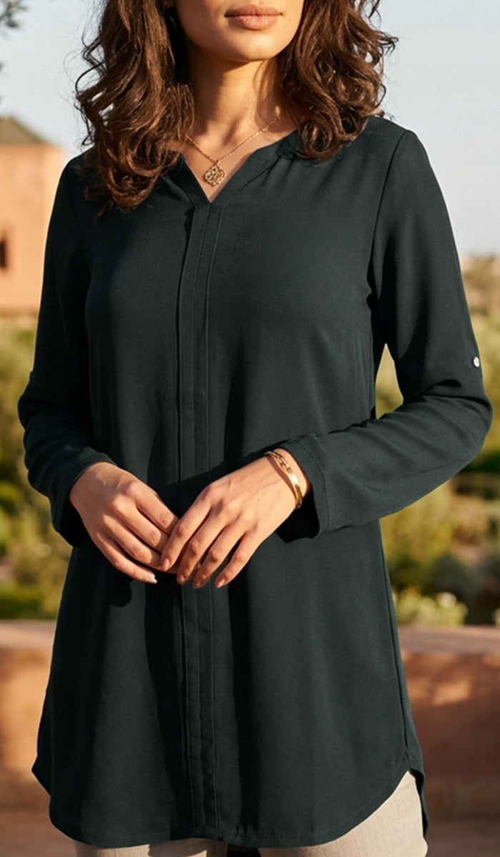 A woman wearing a dark charcoal green long-sleeve button-up tunic top, photographed outdoors with a warm golden-hour background.