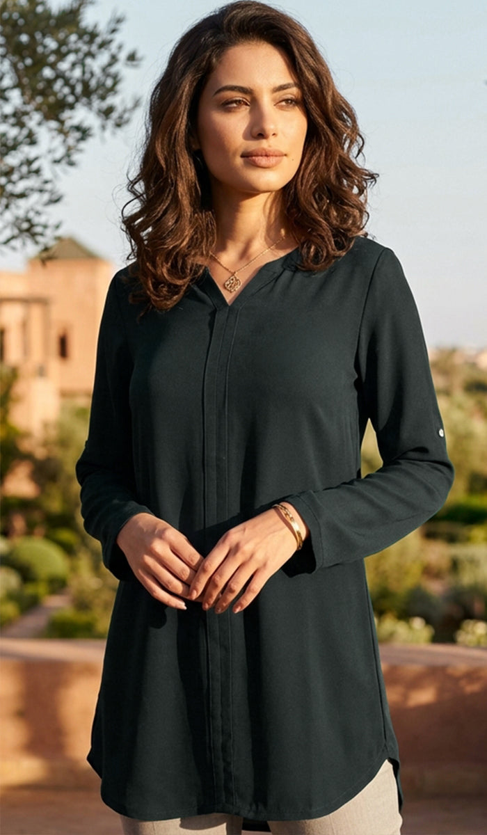 A woman wearing a dark forest green long-sleeve button-up tunic top, standing outdoors with a warm, sunny background.