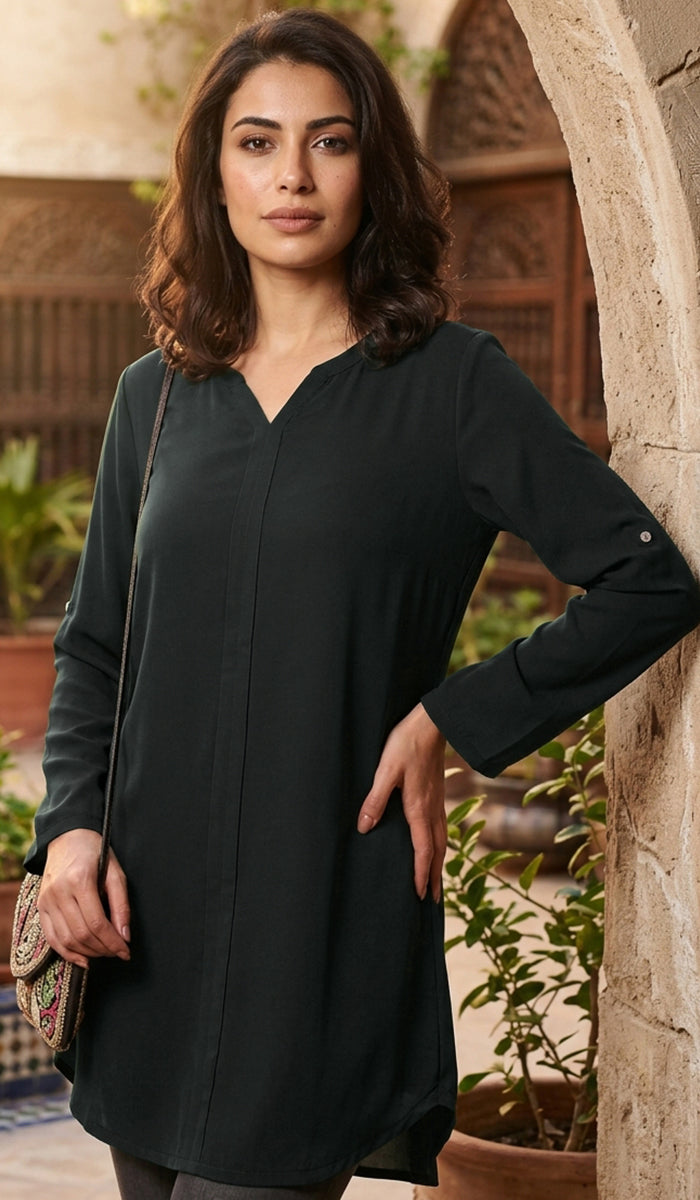A woman wearing a dark teal long-sleeve tunic dress, standing in a courtyard with stone arches and potted plants.
