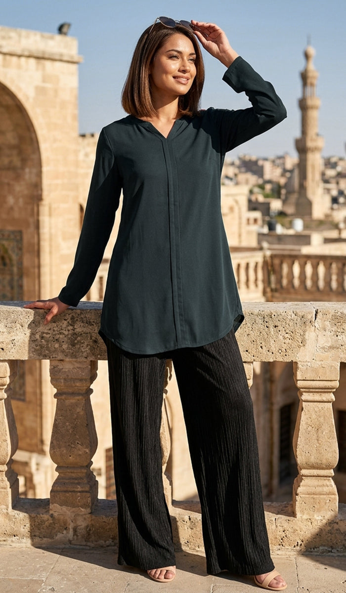 A woman stands outdoors on a stone balcony wearing a dark teal long-sleeve button-front tunic top and black wide-leg pants, with a cityscape and minarets in the background.