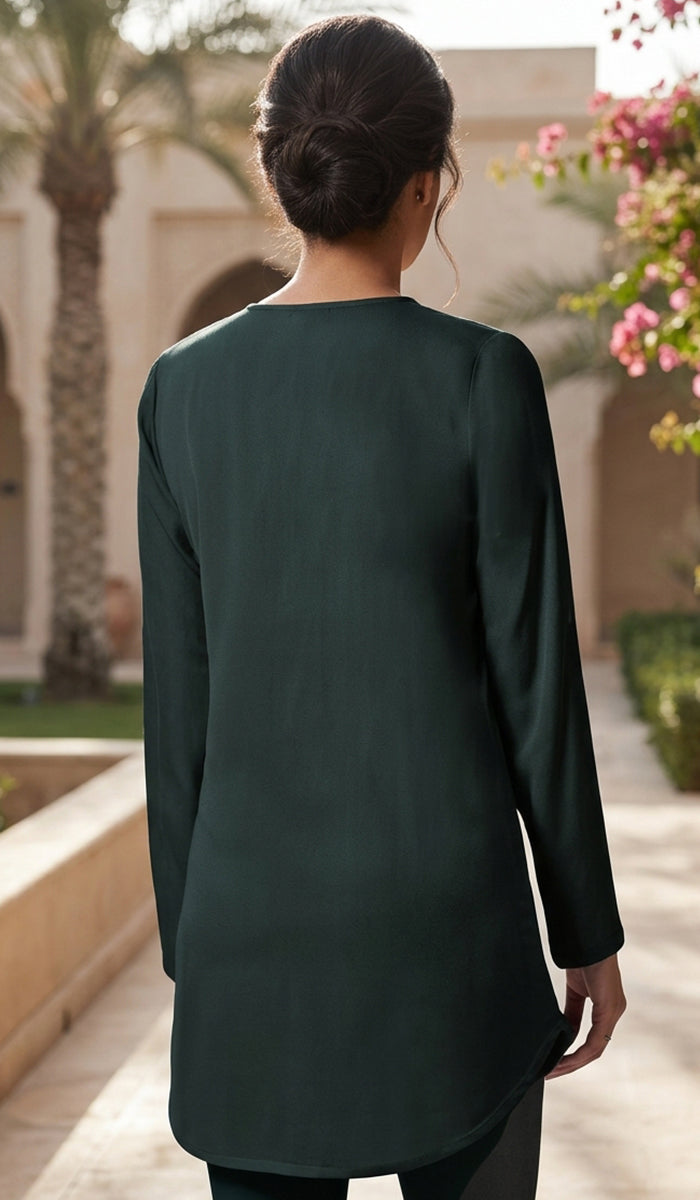 A woman viewed from behind wearing a dark forest green long-sleeve tunic top in an outdoor courtyard setting.