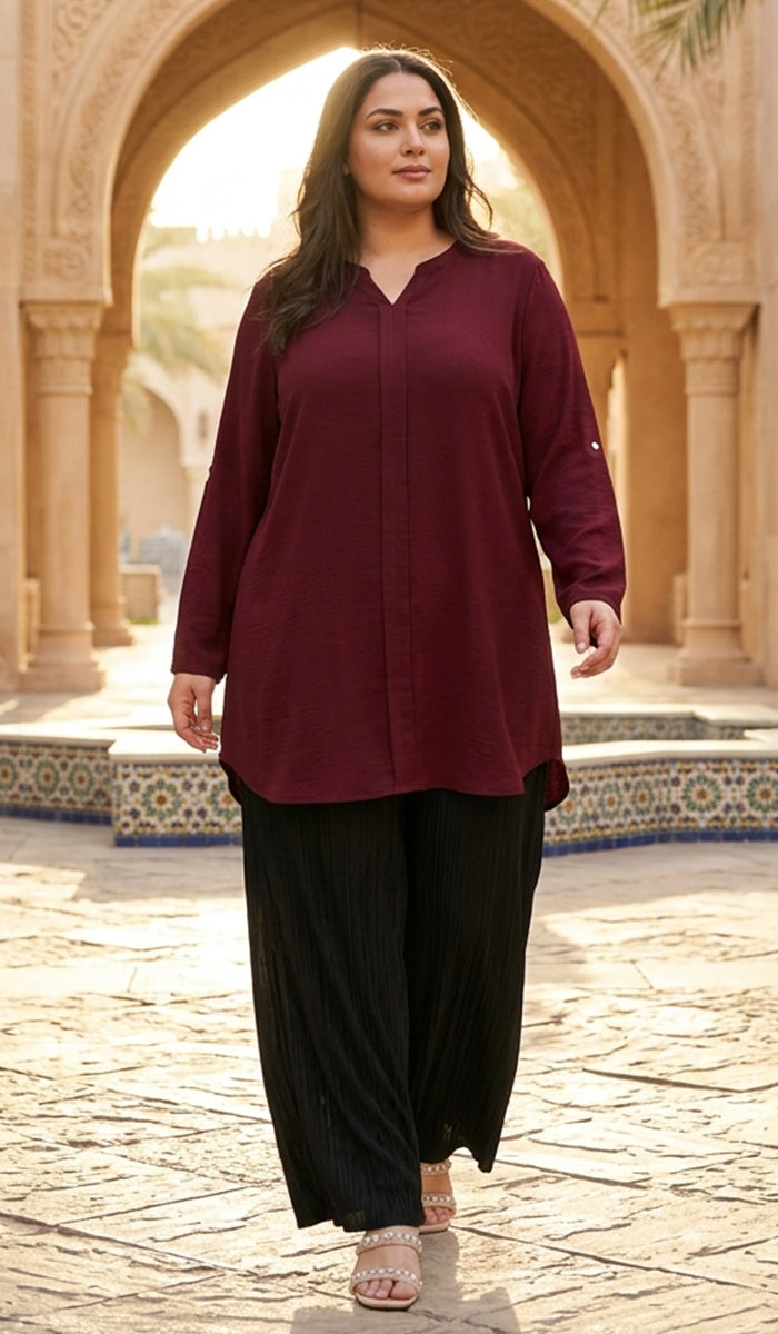 A woman modeling a long-sleeve maroon button-up tunic top with black wide-leg pants, walking through a sunlit stone courtyard framed by arches.