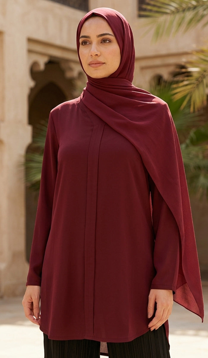 A woman wearing a long-sleeved, draped burgundy hijab and matching tunic top, standing outdoors in front of a beige stone building with arches and palm leaves.