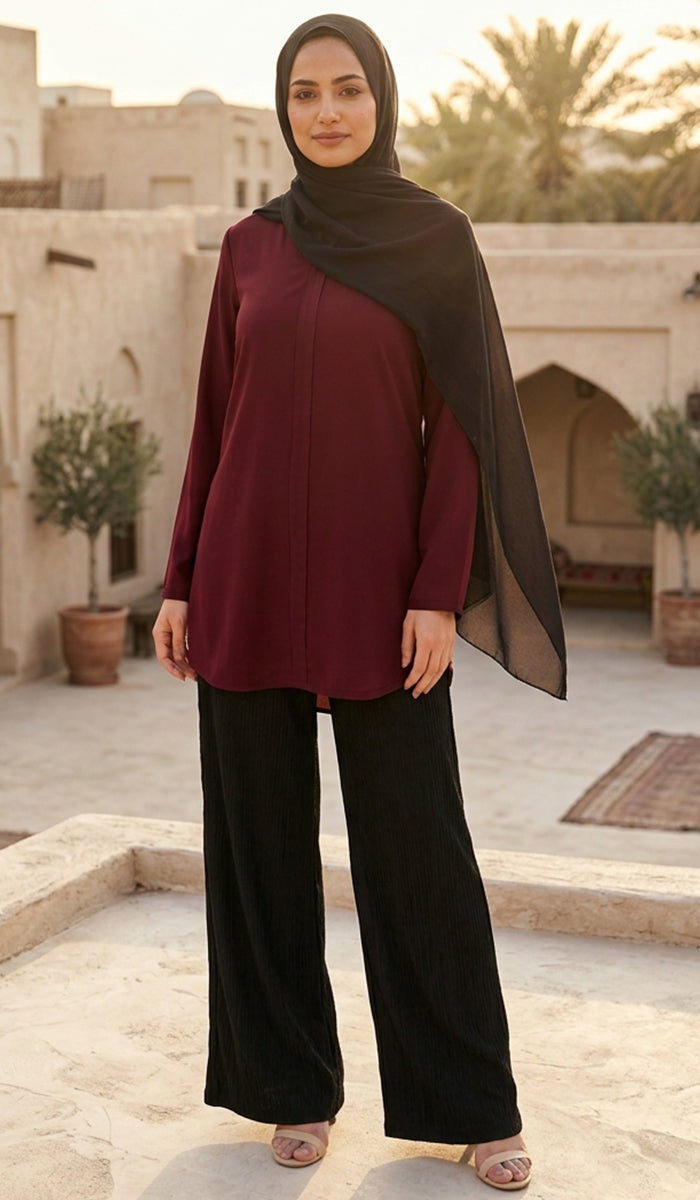 A woman wearing a long-sleeve burgundy abaya with black wide-leg pants and a black hijab, standing in an outdoor courtyard with beige buildings and palm trees in the background.