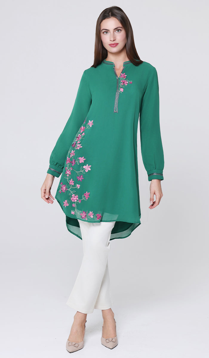 Woman wearing a green embroidered dress with white pants on a light gray background