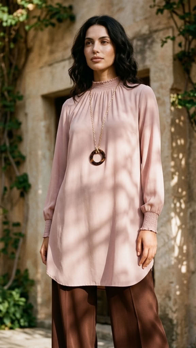 A woman stands outdoors in front of a sunlit stone wall with climbing greenery, wearing a dusty pink long-sleeve tunic top with a gathered neckline and brown wide-leg pants.
