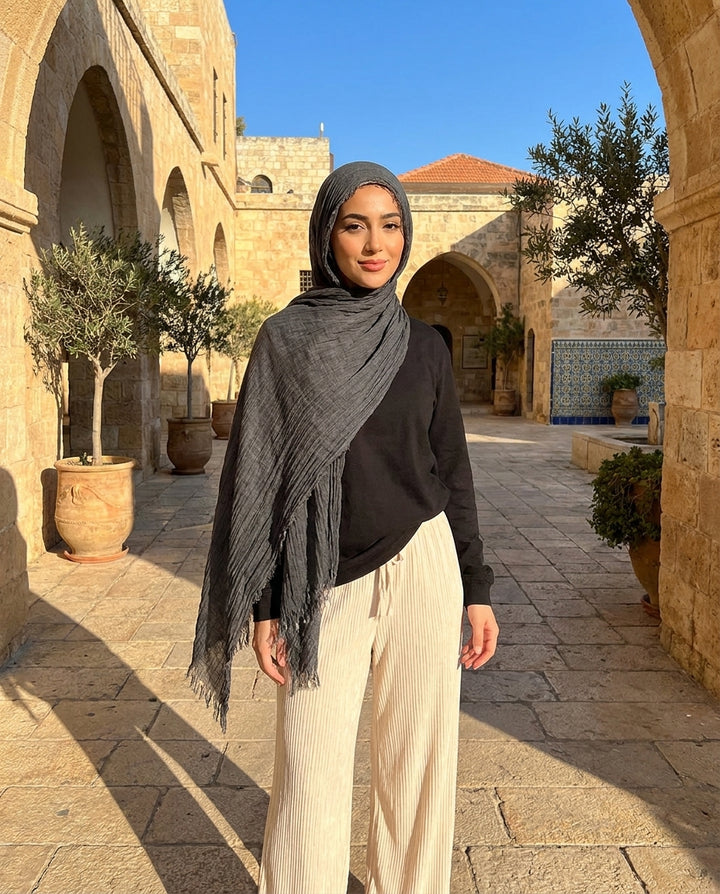 Woman in hijab standing in a sunlit courtyard with stone architecture and potted plants.