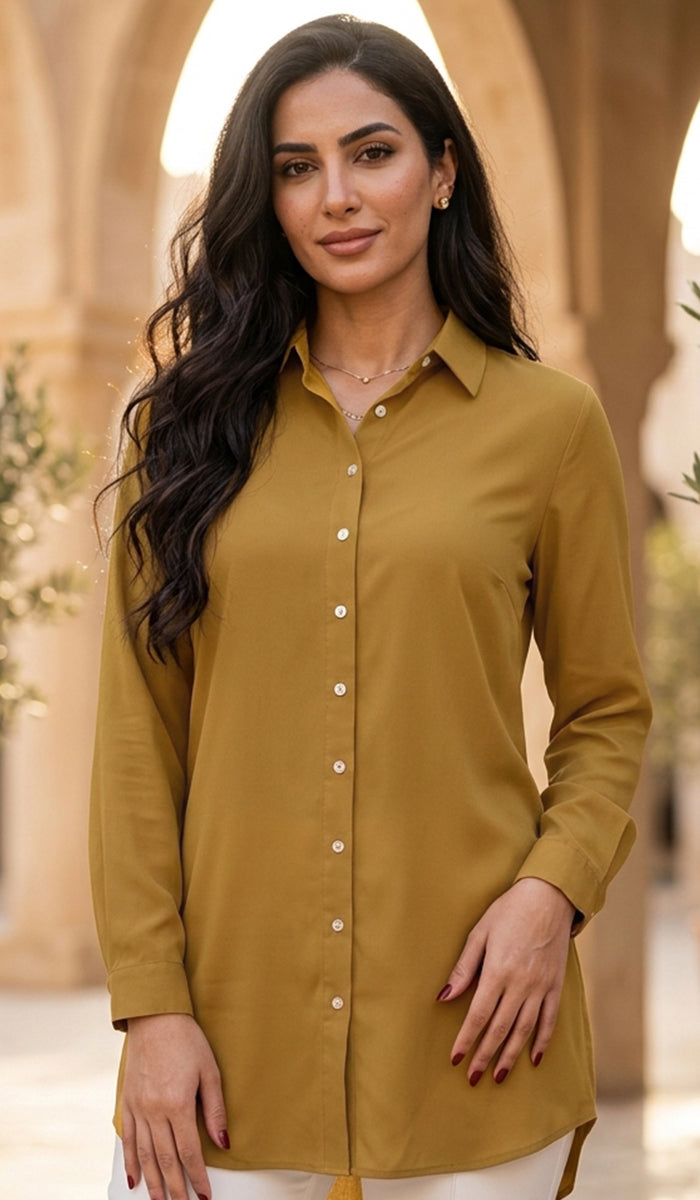 A woman wearing a mustard yellow button-up shirt tunic, standing in front of a blurred beige stone archway background.