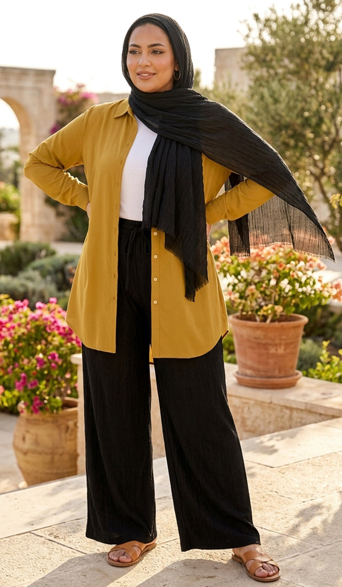 A woman stands outdoors wearing a mustard yellow button-up shirt, black wide-leg pants, a black scarf, and a black head covering. The background shows a garden with potted flowers and stone architecture.