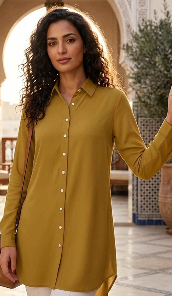A woman wearing a long-sleeve mustard yellow button-up shirt tunic, standing in a sunlit courtyard with arches and tiled walls in the background.