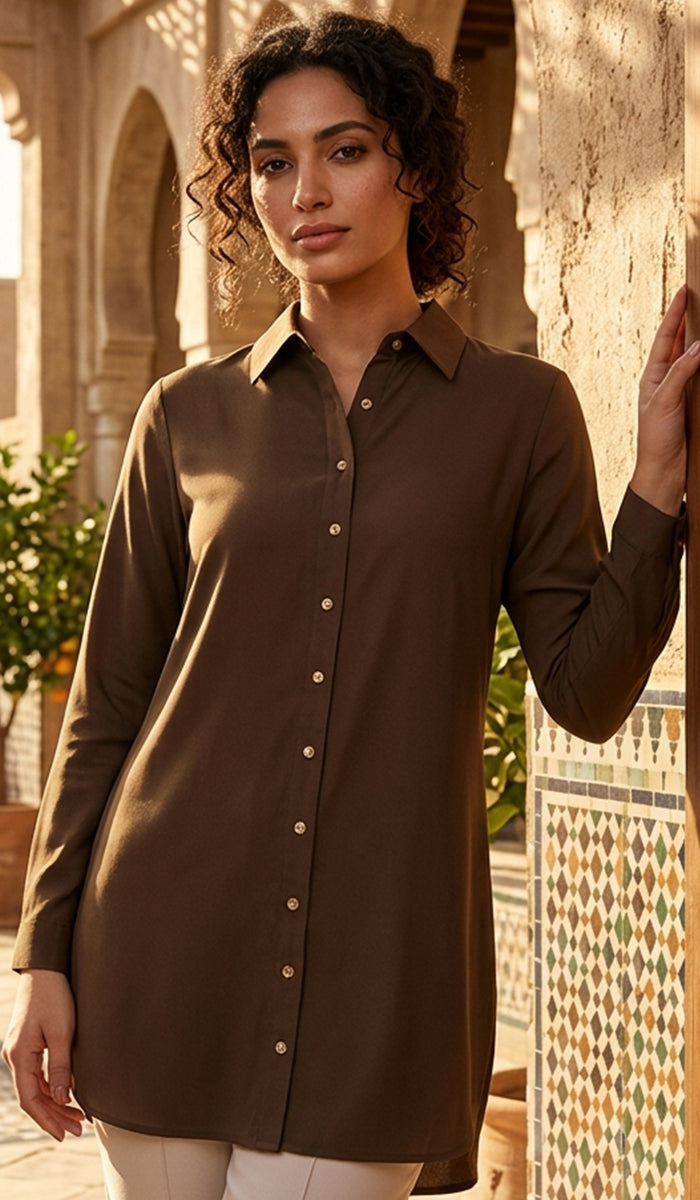 A woman wearing a long-sleeve button-front shirt dress in a warm brown shade, standing in an outdoor architectural setting with stone walls and patterned tile.