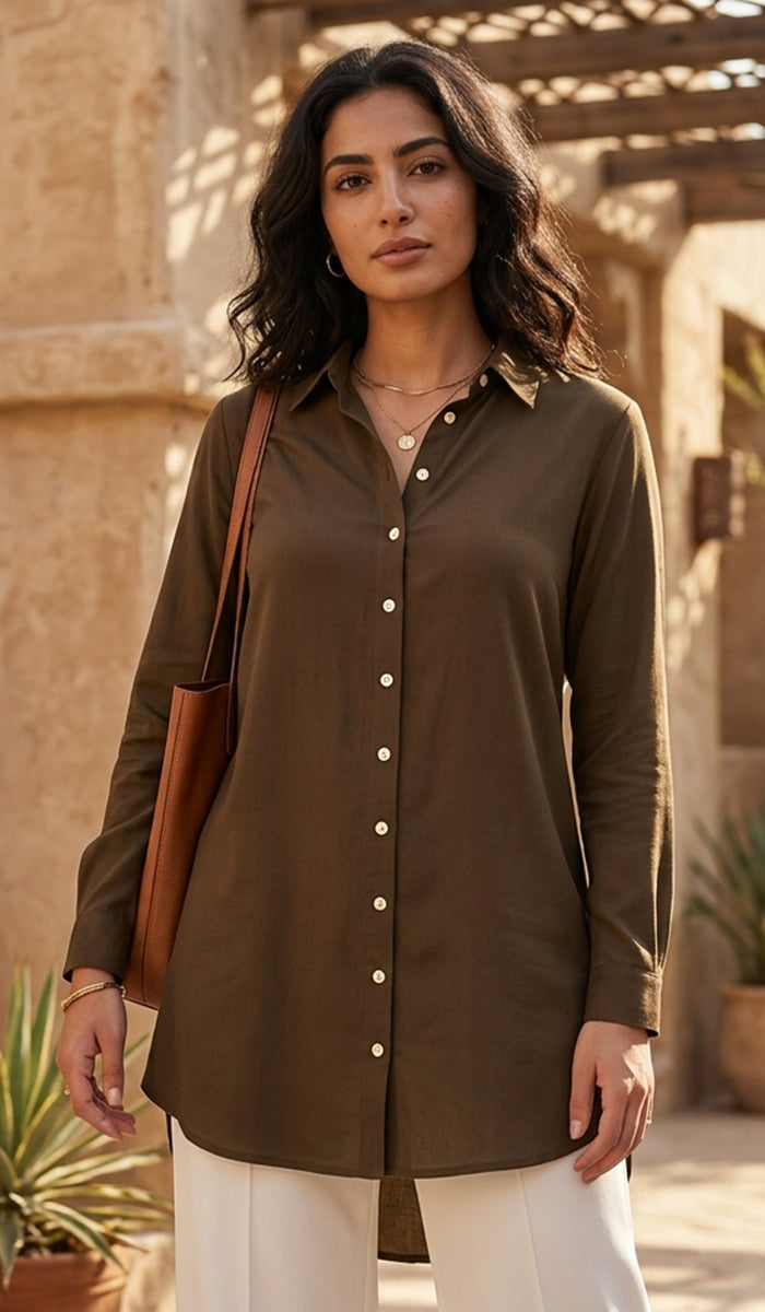 A woman wearing a long-sleeve button-up shirt tunic in a dark olive brown, styled with white pants and a brown leather shoulder bag, set against a warm, sunlit stone patio background.