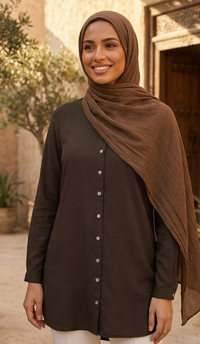 A woman wearing a dark brown button-front tunic and a matching brown hijab, standing outdoors in front of a warm-toned building with stone walls and a wooden door.