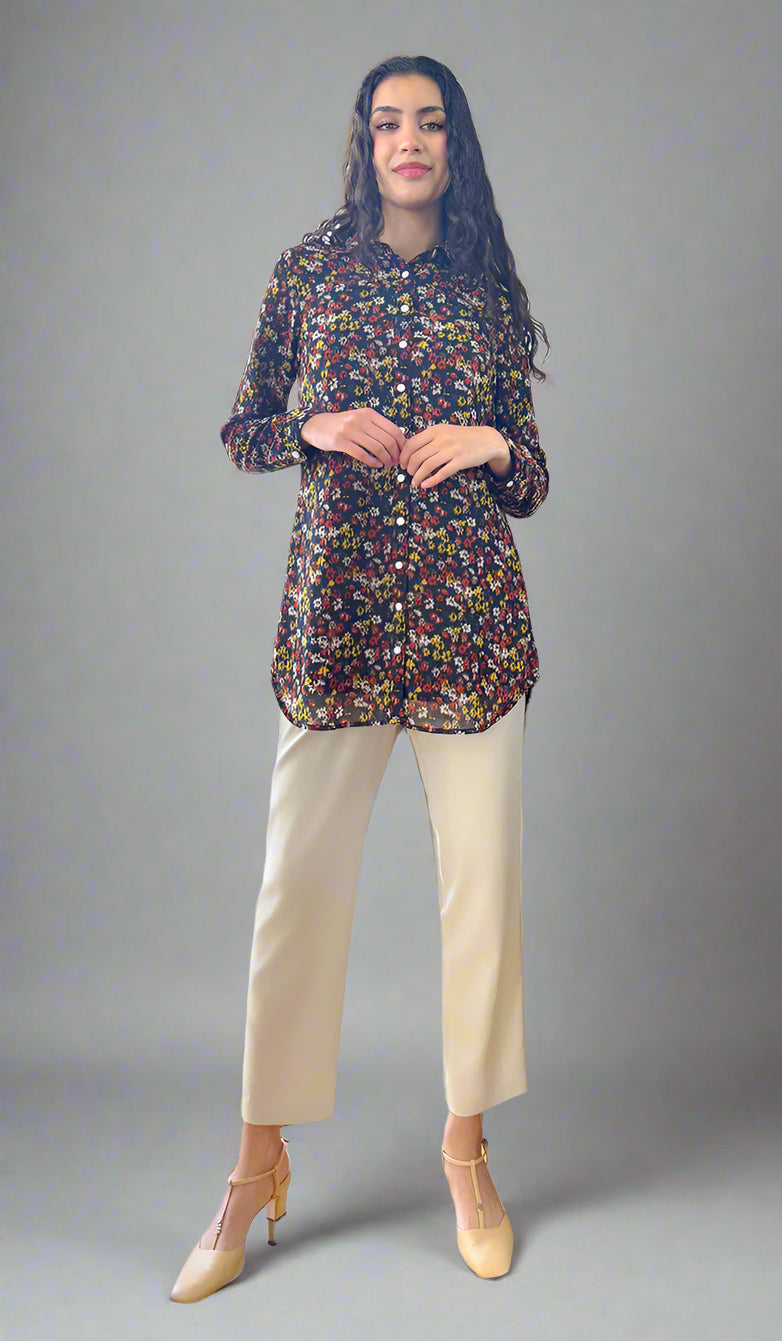 Woman standing in a room with a piano, wearing a floral blouse and white pants.