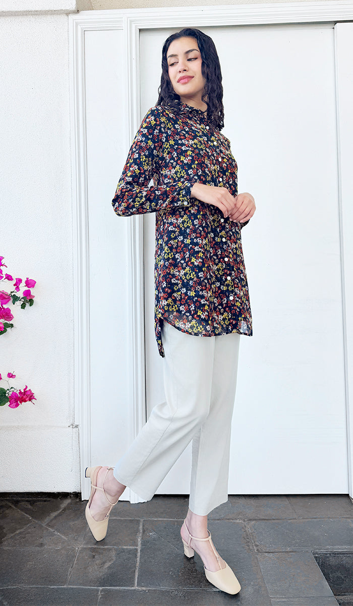Woman wearing a floral blouse and white pants standing in front of a white door.