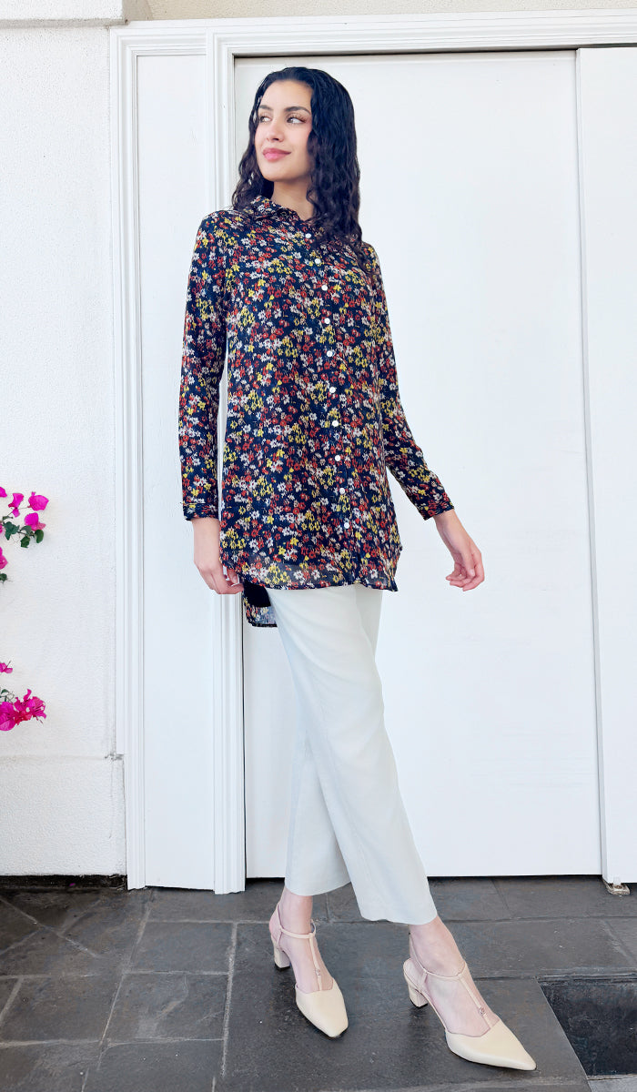 Woman wearing a colorful floral blouse and white pants standing in front of a white door.