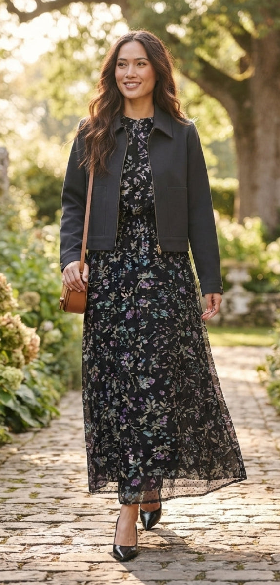 A woman walking on a stone path in a garden, wearing a black floral midi dress with a black cropped jacket, a small brown shoulder bag, and black pointed-toe heels.