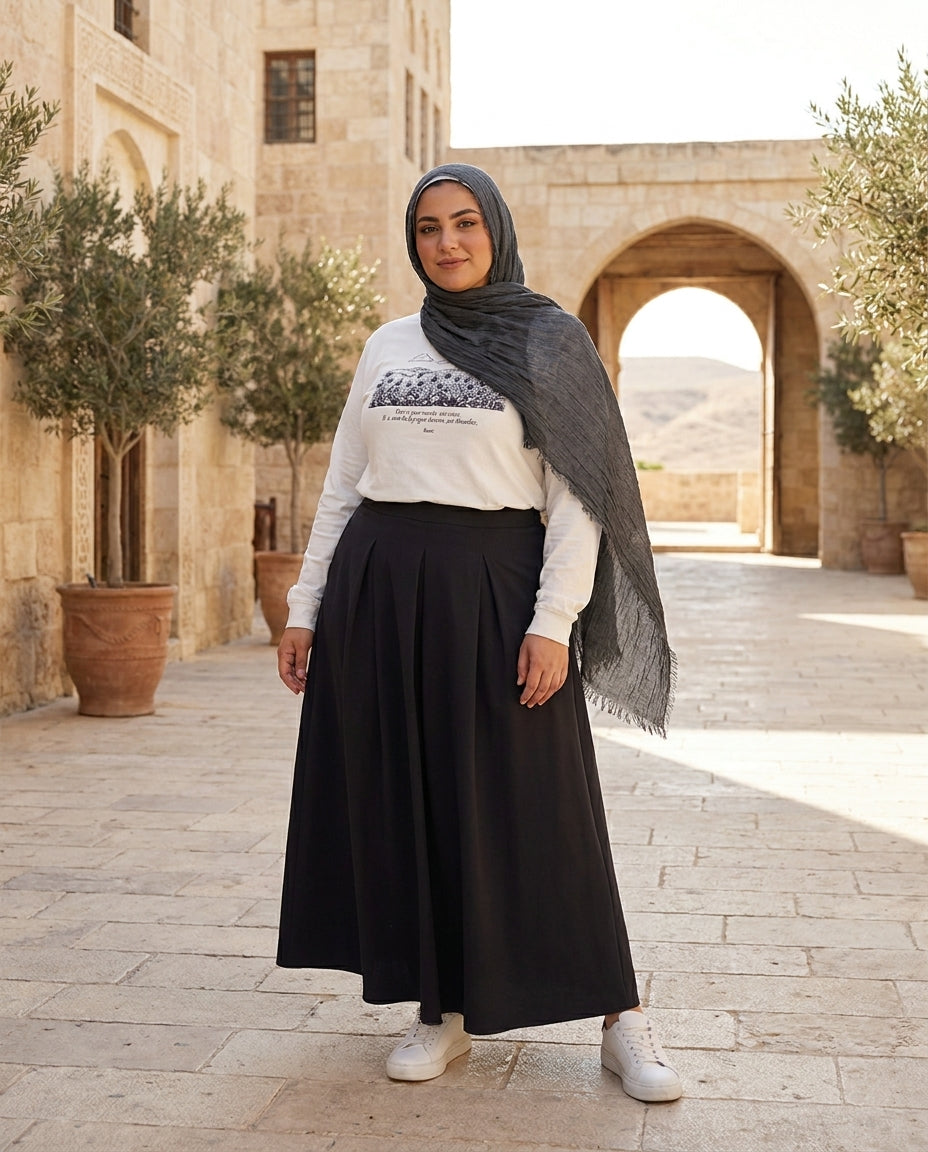 Woman wearing a hijab, white sweater with graphic design, and black skirt standing in front of a stone building with arched doorways.