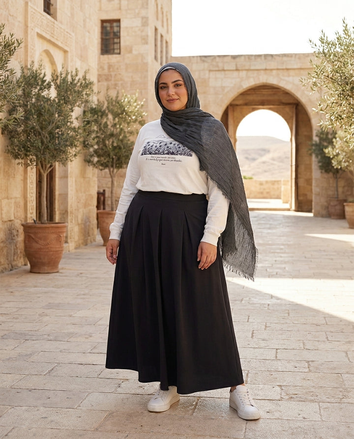 Woman wearing a hijab, white sweater with graphic design, and black skirt standing in front of a stone building with arched doorways.