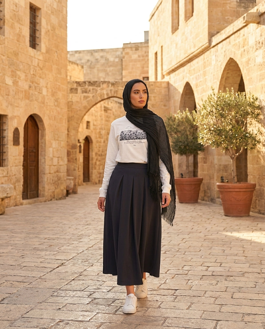 Woman in a hijab standing in an outdoor setting with stone buildings and potted plants.