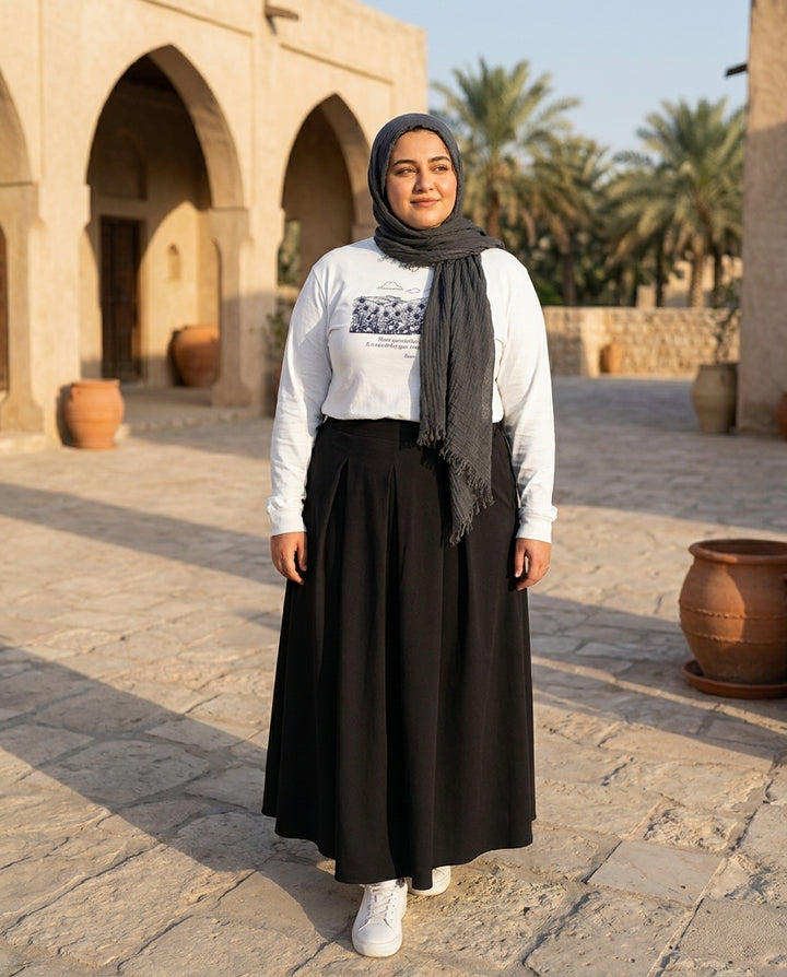 Woman in a black abaya and white shirt standing in an outdoor setting with arches and palm trees.