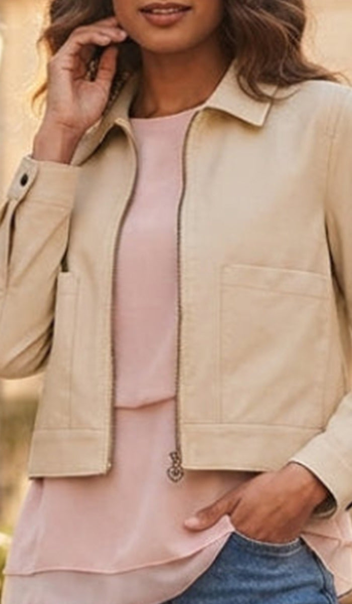 A woman wearing a light beige zip-front jacket over a pale pink top and blue jeans, shown in a softly blurred outdoor setting.