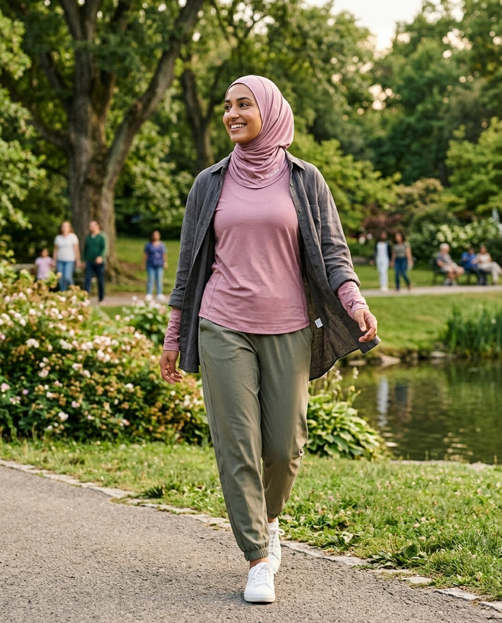 Woman walking in a park wearing a pink hijab, pink shirt, and green pants.