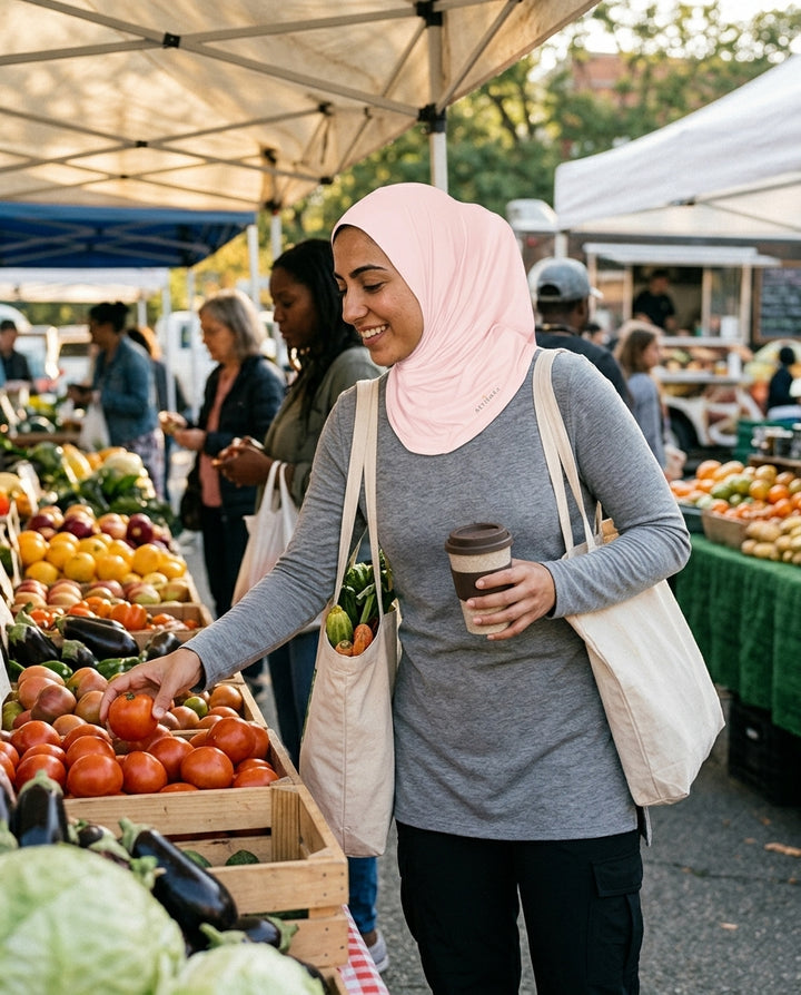 Woman shopping at an outdoor farmers market, holding a coffee cup and a reusable bag.