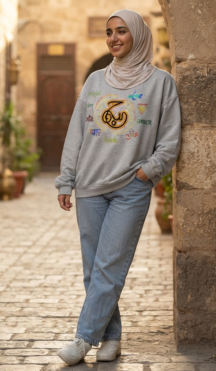 A woman wearing a light gray sweatshirt with colorful Arabic calligraphy and a crescent moon graphic, paired with light blue jeans and a beige hijab, standing on a cobblestone street.