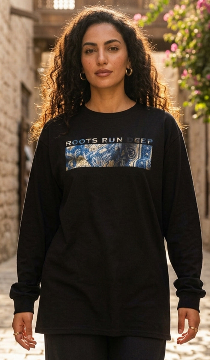A woman wearing a black long-sleeve graphic t-shirt with 'ROOTS RUN DEEP' and a blue abstract graphic on the chest, standing in an outdoor stone alleyway.