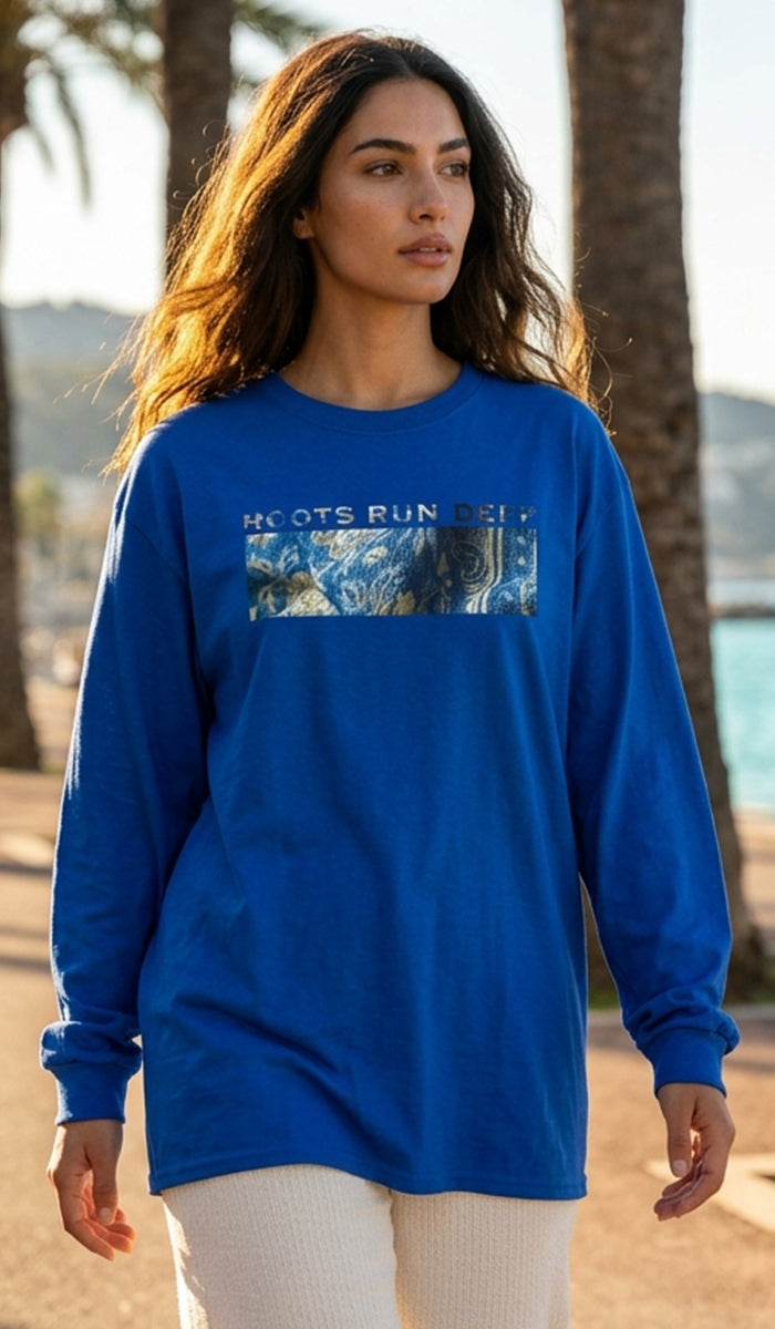 A woman wearing a royal blue long-sleeve t-shirt with a graphic on the chest that reads 'ROOTS RUN DEEP' above a scenic landscape print, standing outdoors near palm trees and water.
