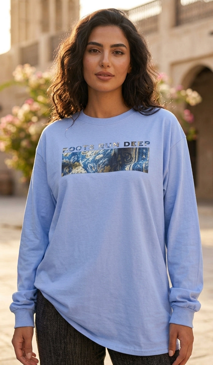 A woman wearing a light blue long-sleeve t-shirt with a graphic print on the chest that reads 'ROOTS RUN DEEP' above a scenic landscape image, set against an outdoor background with blurred buildings and flowers.