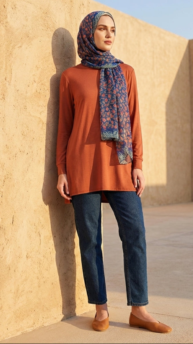 Woman wearing an orange top and blue jeans with a patterned scarf, standing against a beige wall.