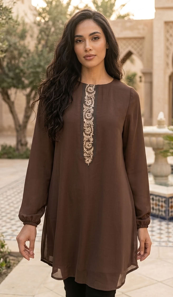 A woman wearing a brown long-sleeve tunic dress with intricate gold embroidery down the center front, standing in an outdoor courtyard setting.