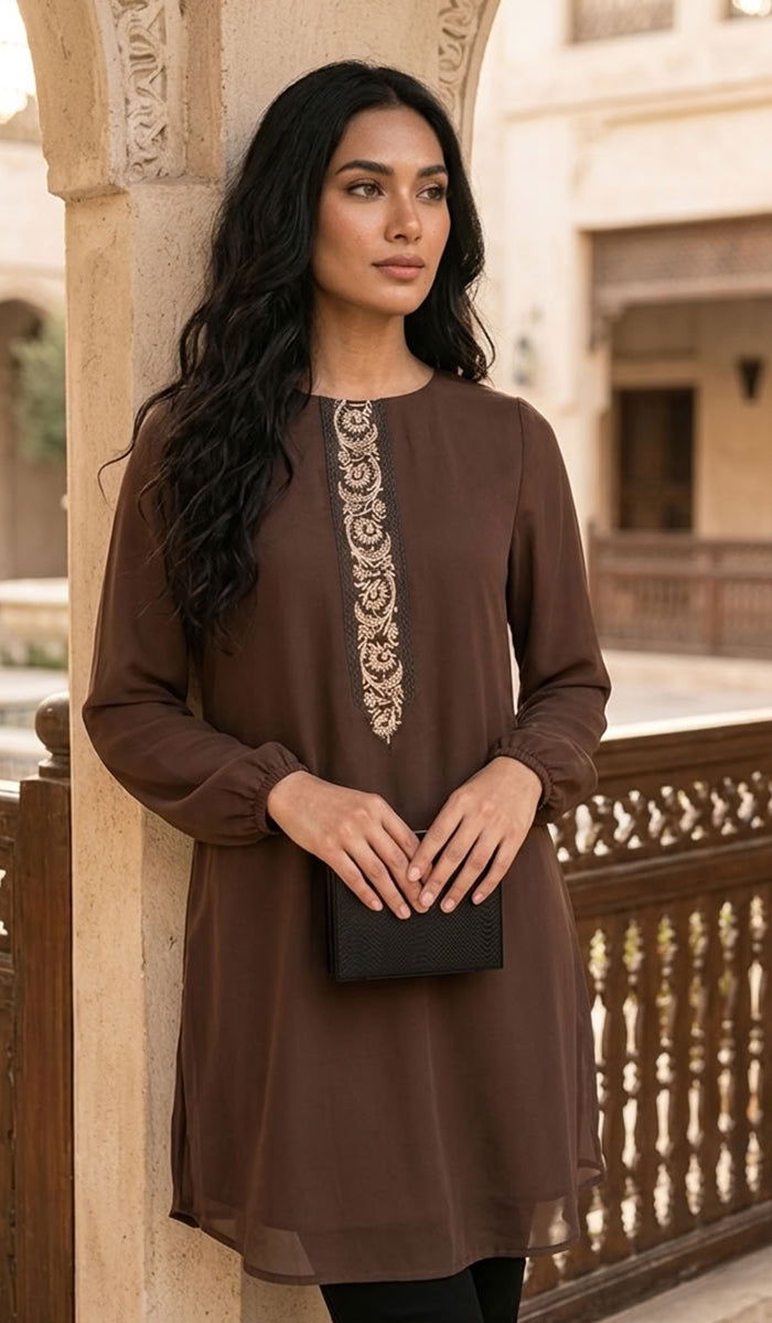 A woman wearing a brown long-sleeve tunic with intricate gold embroidery down the center front, holding a black clutch bag in an outdoor architectural setting.