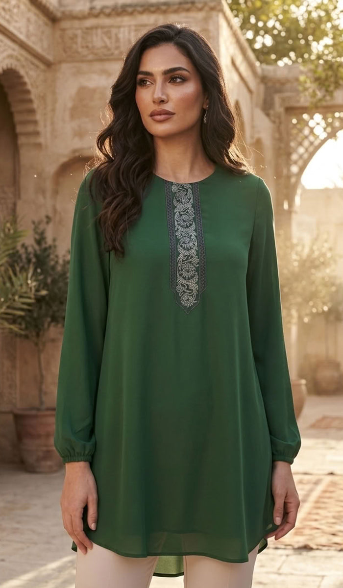 A woman wearing a long-sleeved sage green tunic top with a decorative gray embroidered neckline, standing in front of a warm, sunlit stone courtyard with arches.