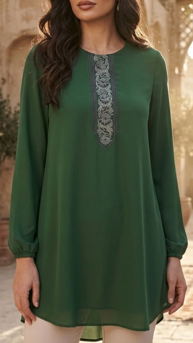 A woman wearing a long-sleeve green tunic top with a decorative embroidered yoke panel, shown from the lips down in an outdoor setting with warm blurred background.