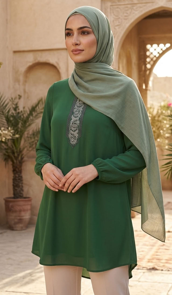 A woman wearing a sage green long-sleeve tunic with a decorative embroidered neckline, paired with a matching sage green hijab and light beige pants, standing in front of a warm-toned architectural background with arches and potted plants.