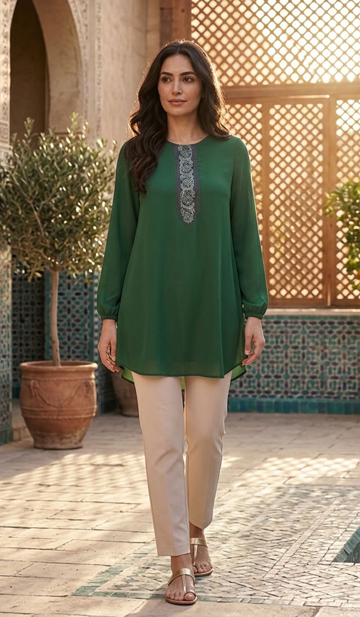 A woman modeling a green long-sleeve kurta with a silver embroidered neckline, paired with beige trousers, standing in a sunlit courtyard with tiled walls and wooden lattice screens.
